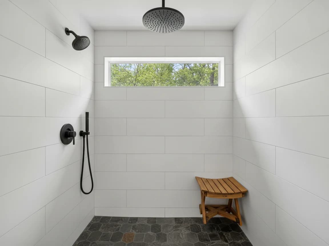 Walk-in shower with white large-format tile, a black rain head and handheld sprayer, a teak stool and a slate-look mosaic floor under a high window