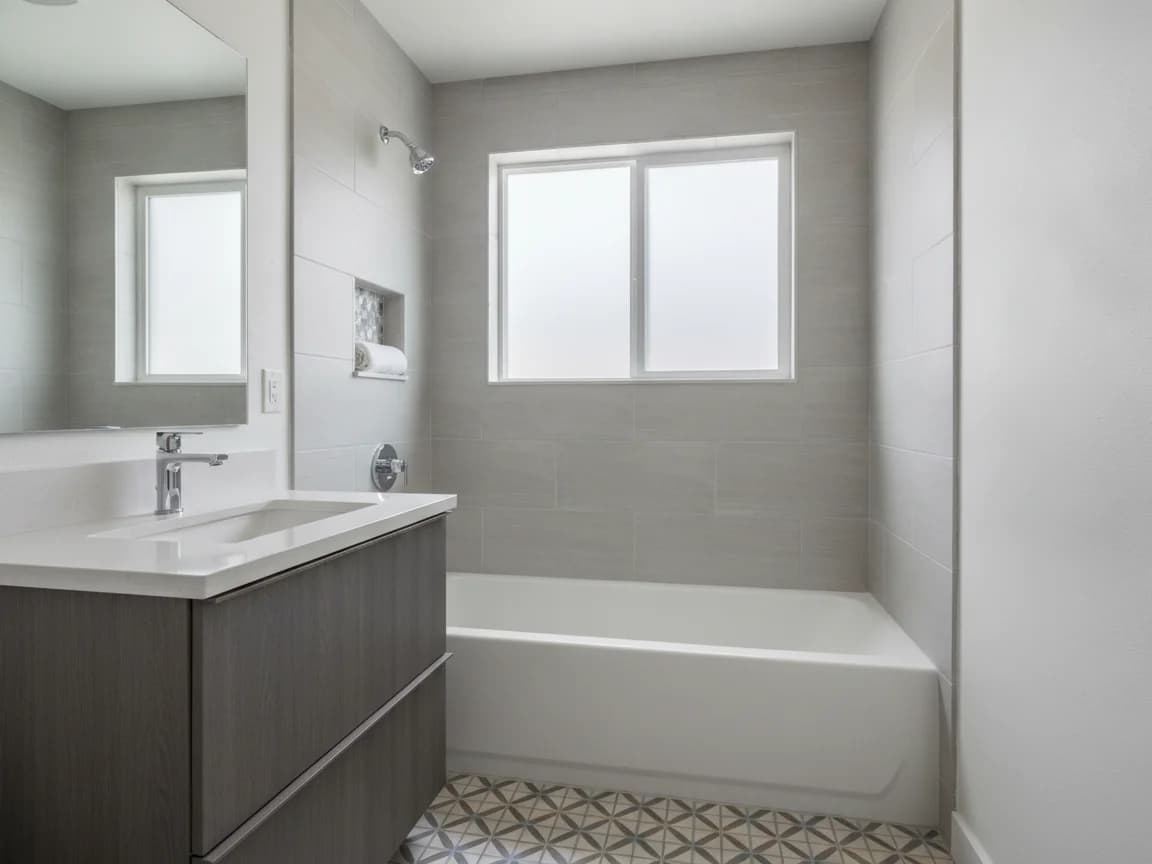 Hall bathroom with a white tub in a gray tile surround with a recessed niche, a gray floating vanity and patterned floor tile