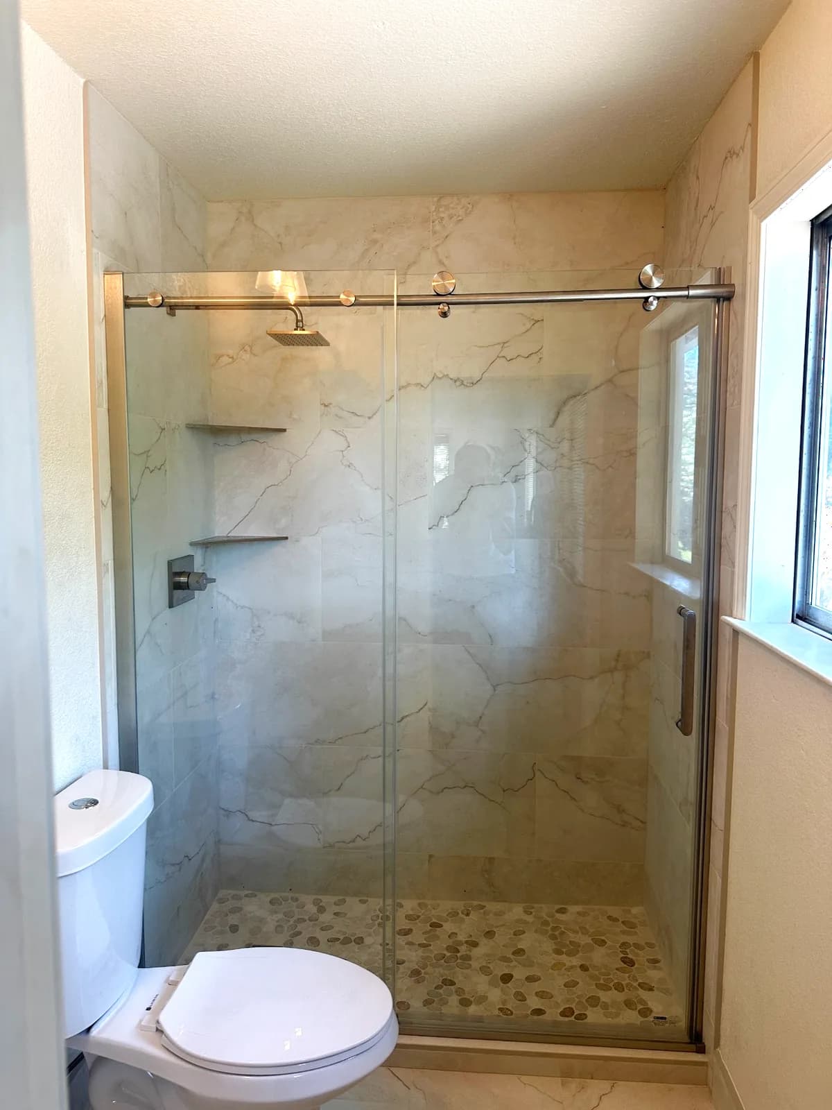 After: walk-in shower with marble-look porcelain, tiled window, pebble floor and frameless sliding glass in Auburn, CA