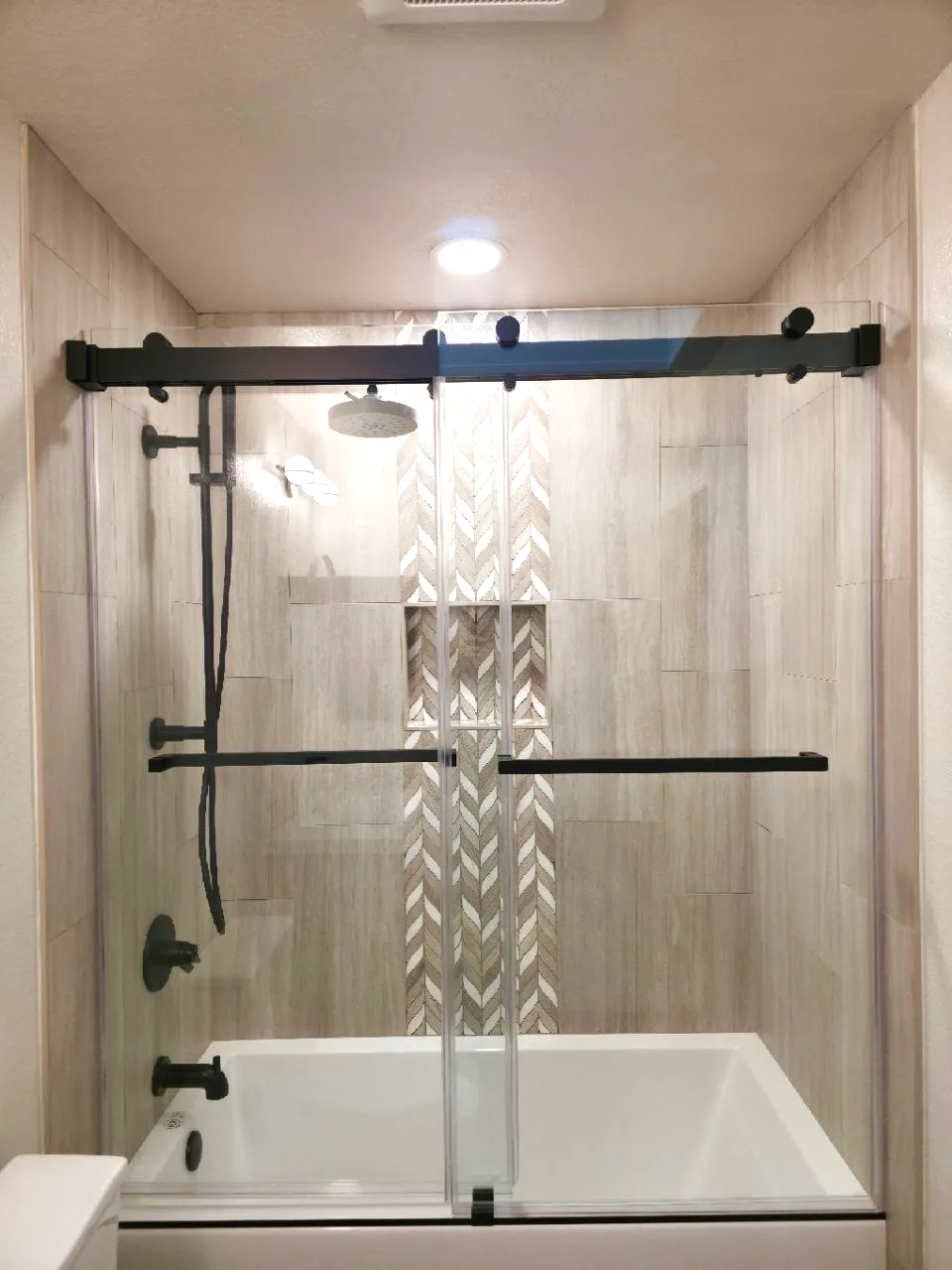 After: alcove tub with wood-look porcelain walls, chevron accent column and matte black sliding glass in Cameron Park, CA