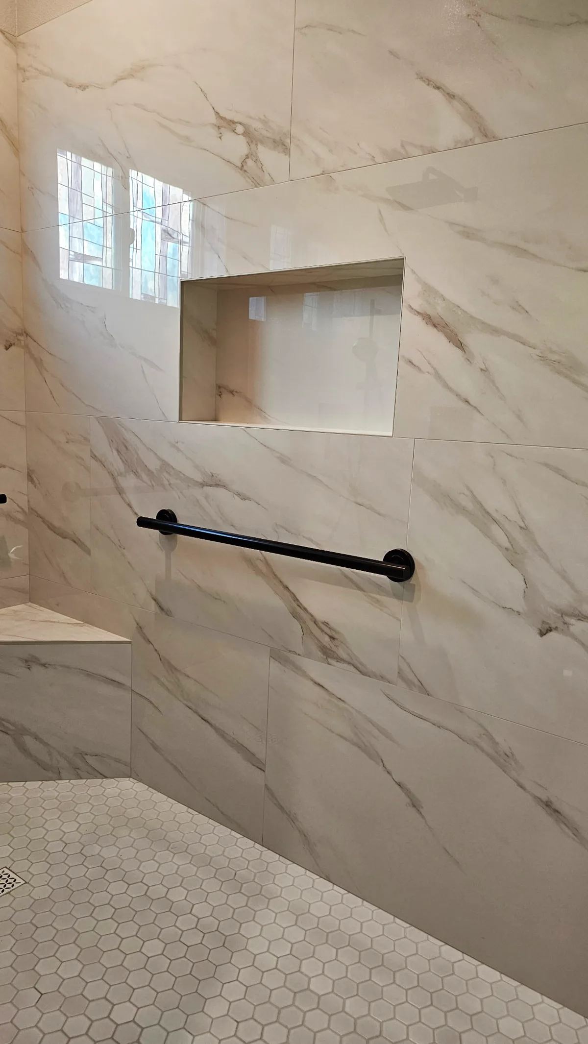After: Roseville wheelchair-accessible roll-in shower with large-format Calacatta marble-look porcelain walls, matte black 36-inch grab bar, recessed niche, white hex mosaic floor and custom marble corner bench