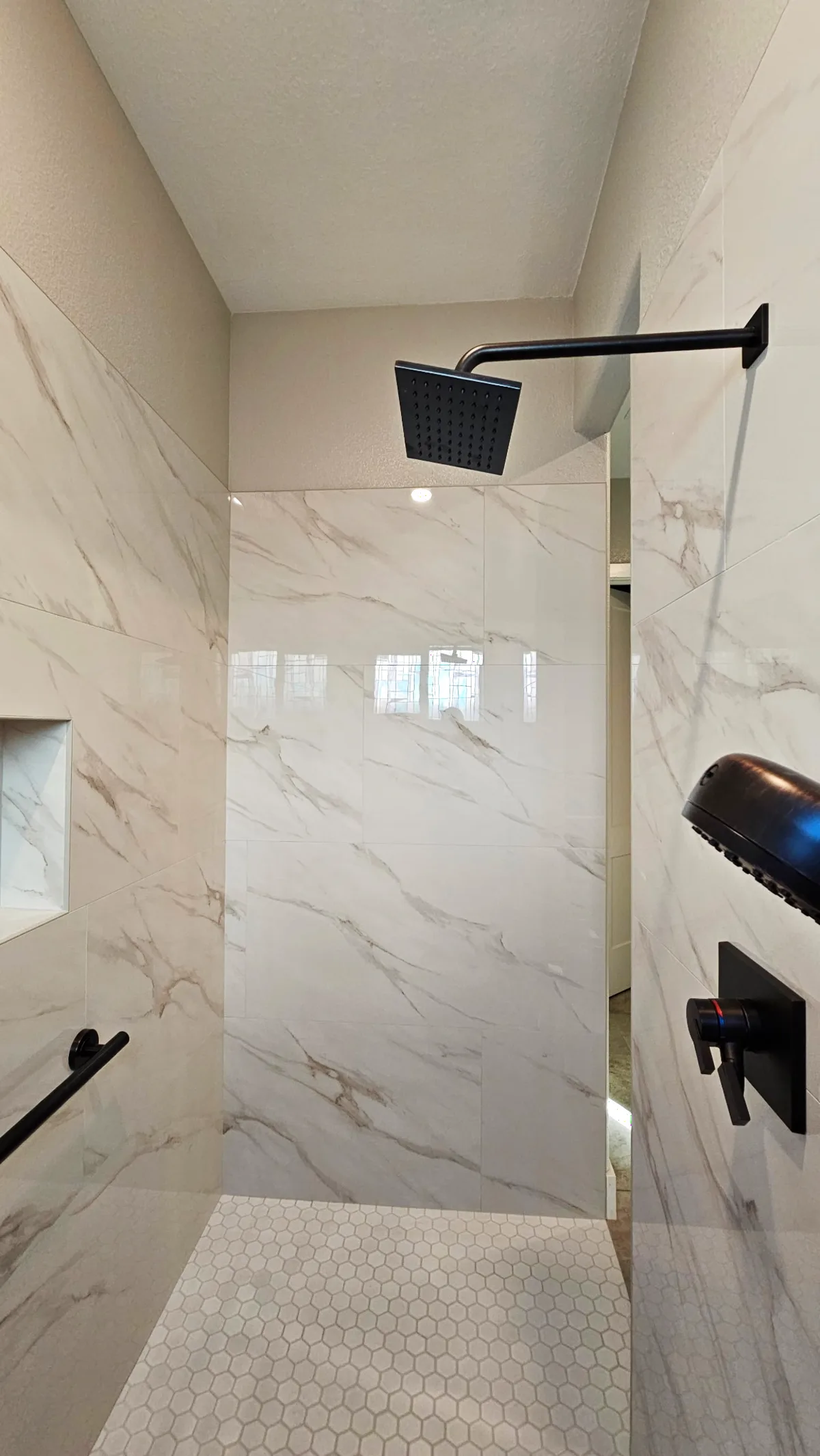 After: Roseville roll-in shower overhead view with matte black square rain head, matte black handheld, dual square controls, Calacatta porcelain walls and white hex mosaic floor at the shower entry