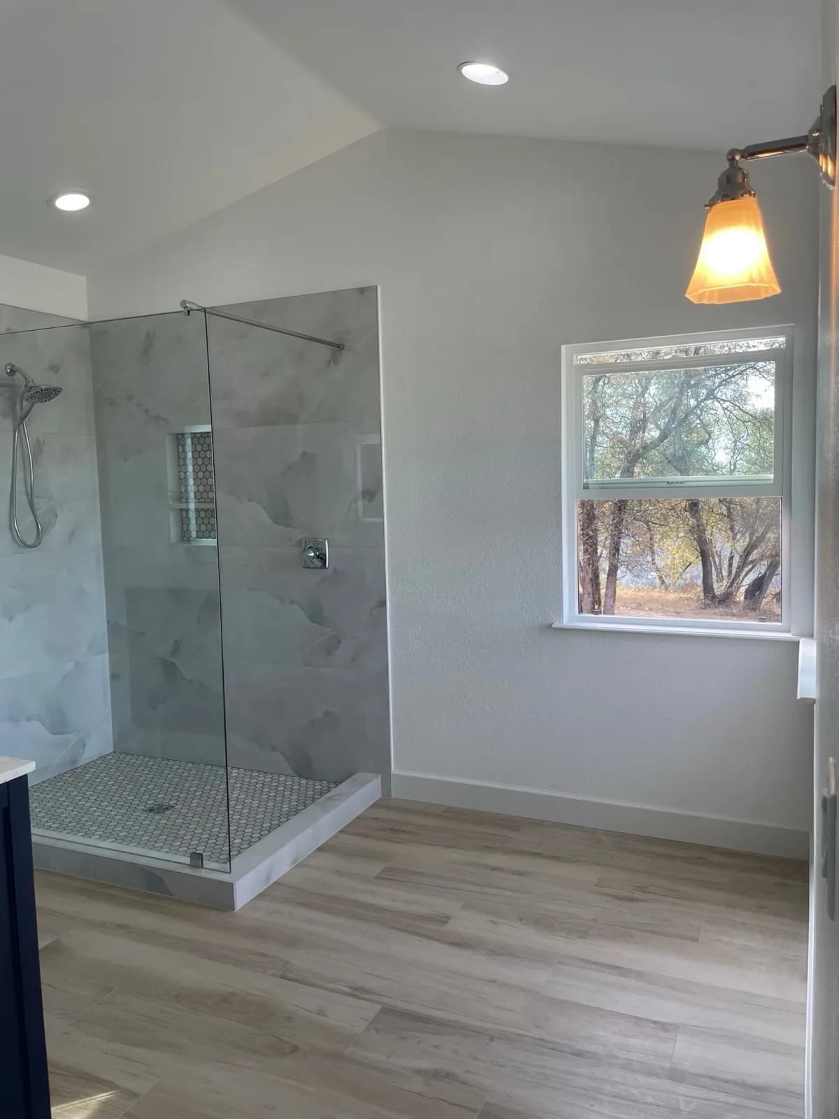 After: Pilot Hill master bathroom with open walk-in marble-look tile shower, frameless glass panel, hex mosaic base and wood-look plank floor