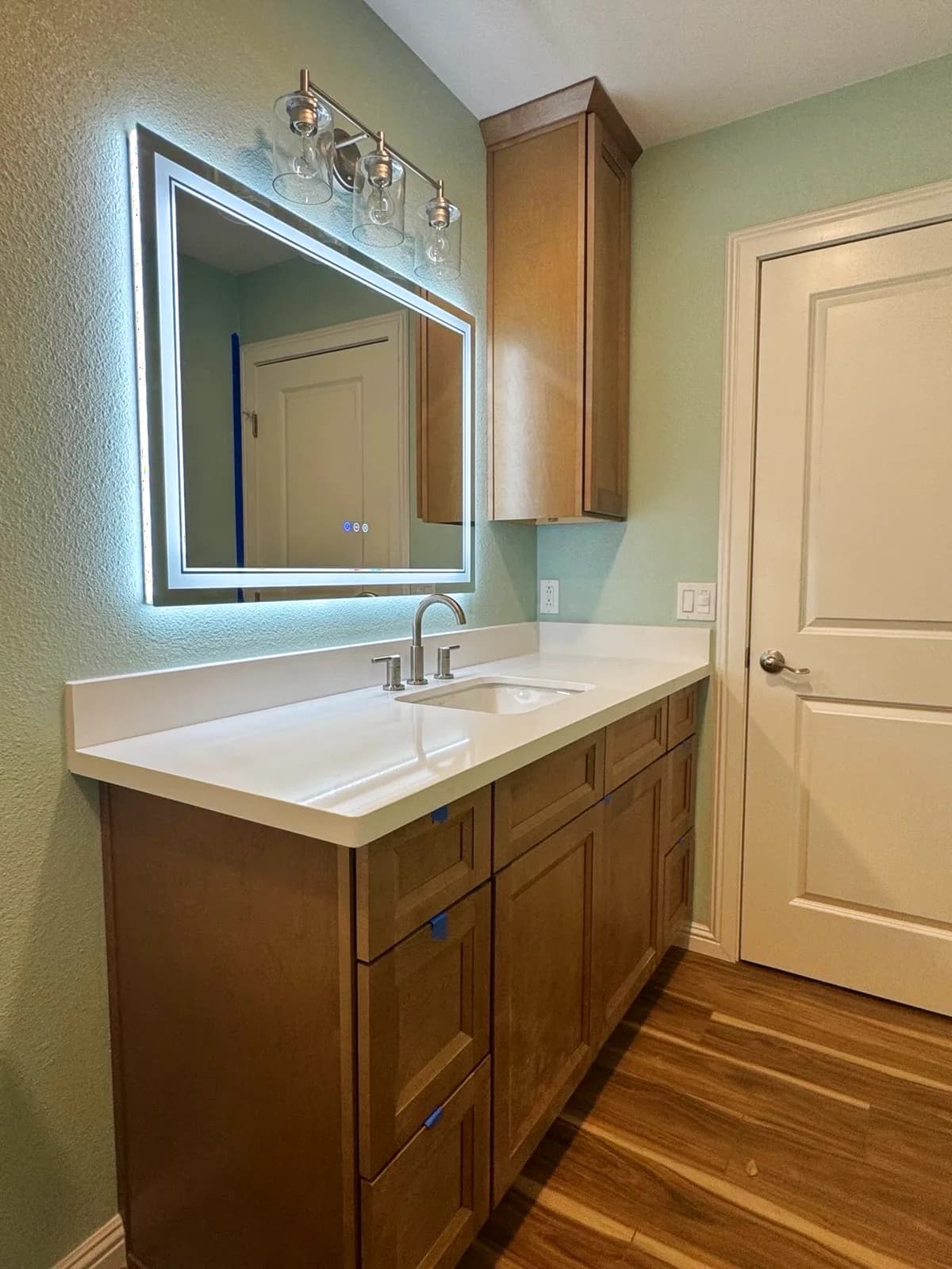 After: Roseville bathroom warm-wood shaker vanity with white quartz top, backlit LED mirror, chrome faucet, upper wall cabinet and wood-look plank flooring