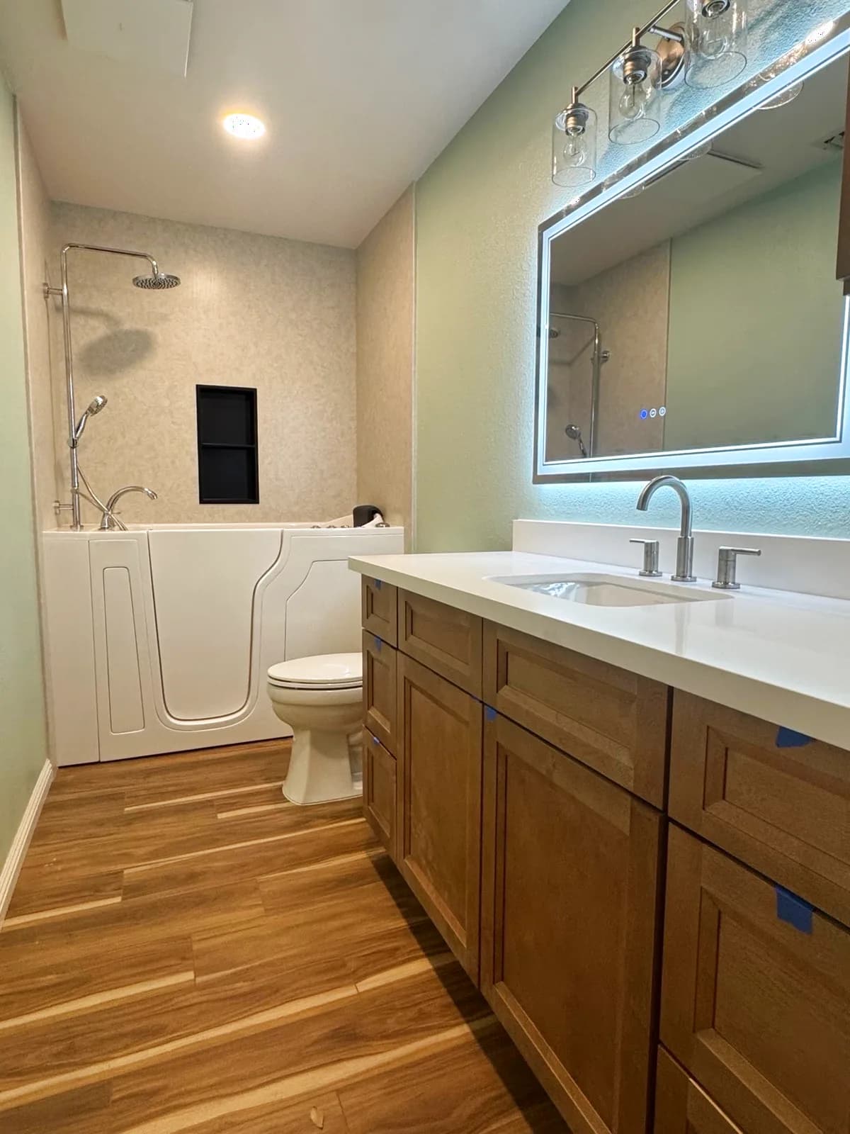 After: Roseville bathroom shaker vanity, backlit LED mirror and walk-in tub with wood-look plank floor running from the vanity to the tub alcove