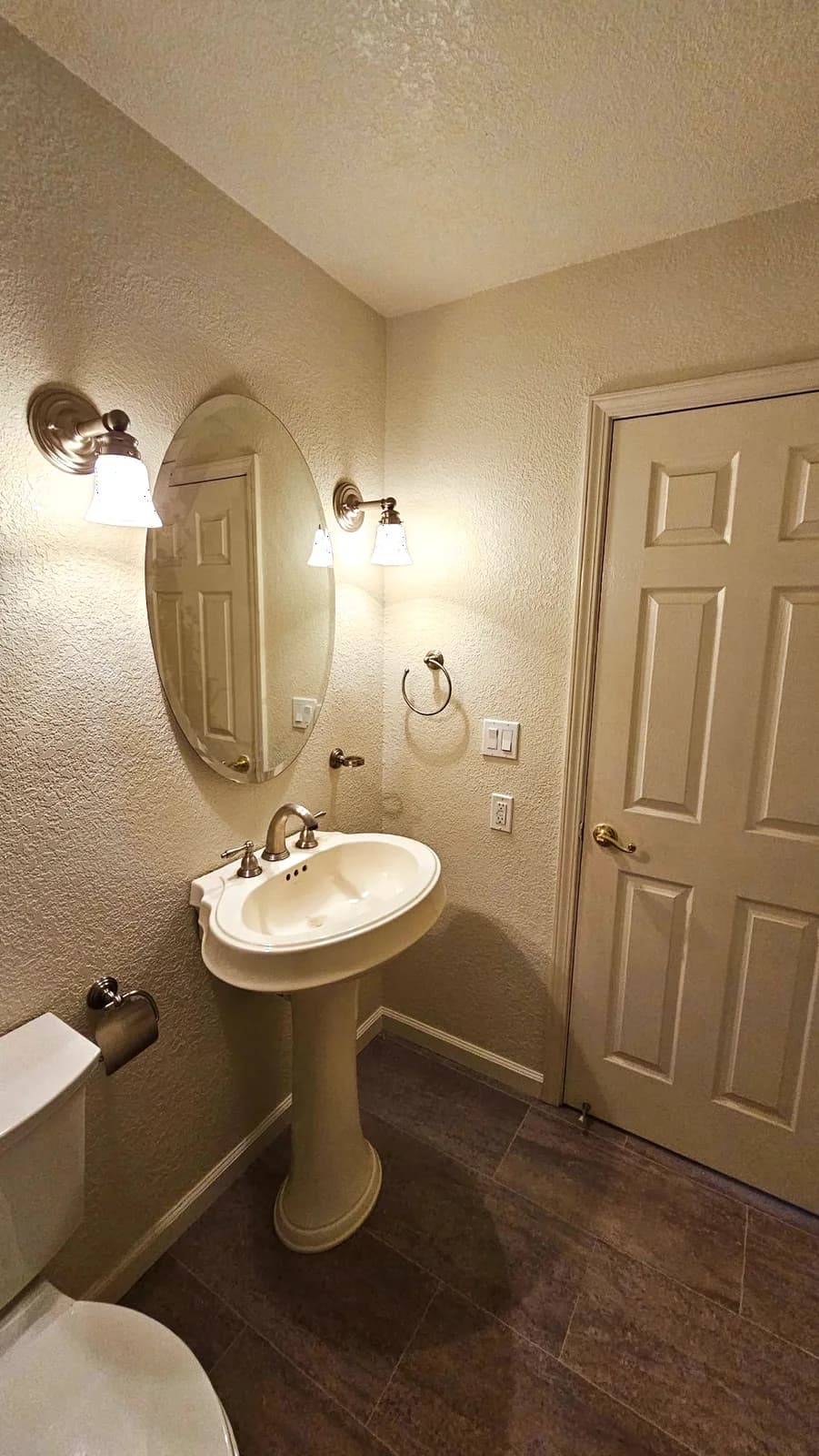 After: pedestal sink, wall sconces and oval mirror above new vinyl plank flooring in a Woodland hallway bathroom